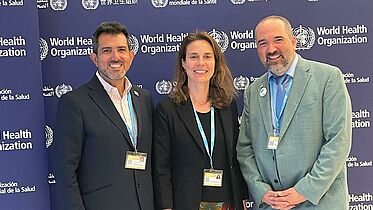 Three CBM representatives stand together, smiling, in front of a backdrop displaying the World Health Organization logo. They are attending the 77th World Health Assembly, showcasing CBM's commitment to inclusive health initiatives and global health policy advocacy.